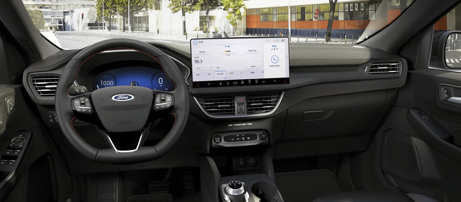 Interior of a modern vehicle with a steering wheel, dashboard, and a large touchscreen display in the center console, with a city street background visible through the windshield.