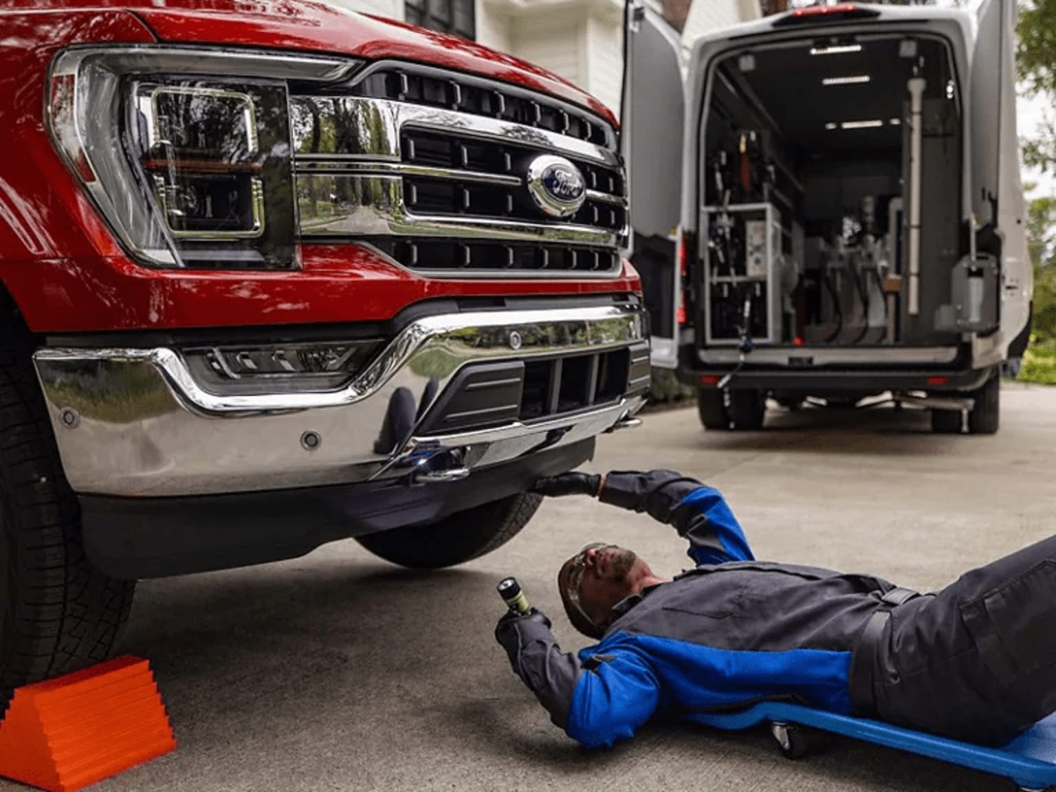 A mechanic in a blue uniform is working on the underside of a large red truck in a garage setting, with another vehicle visible in the background.