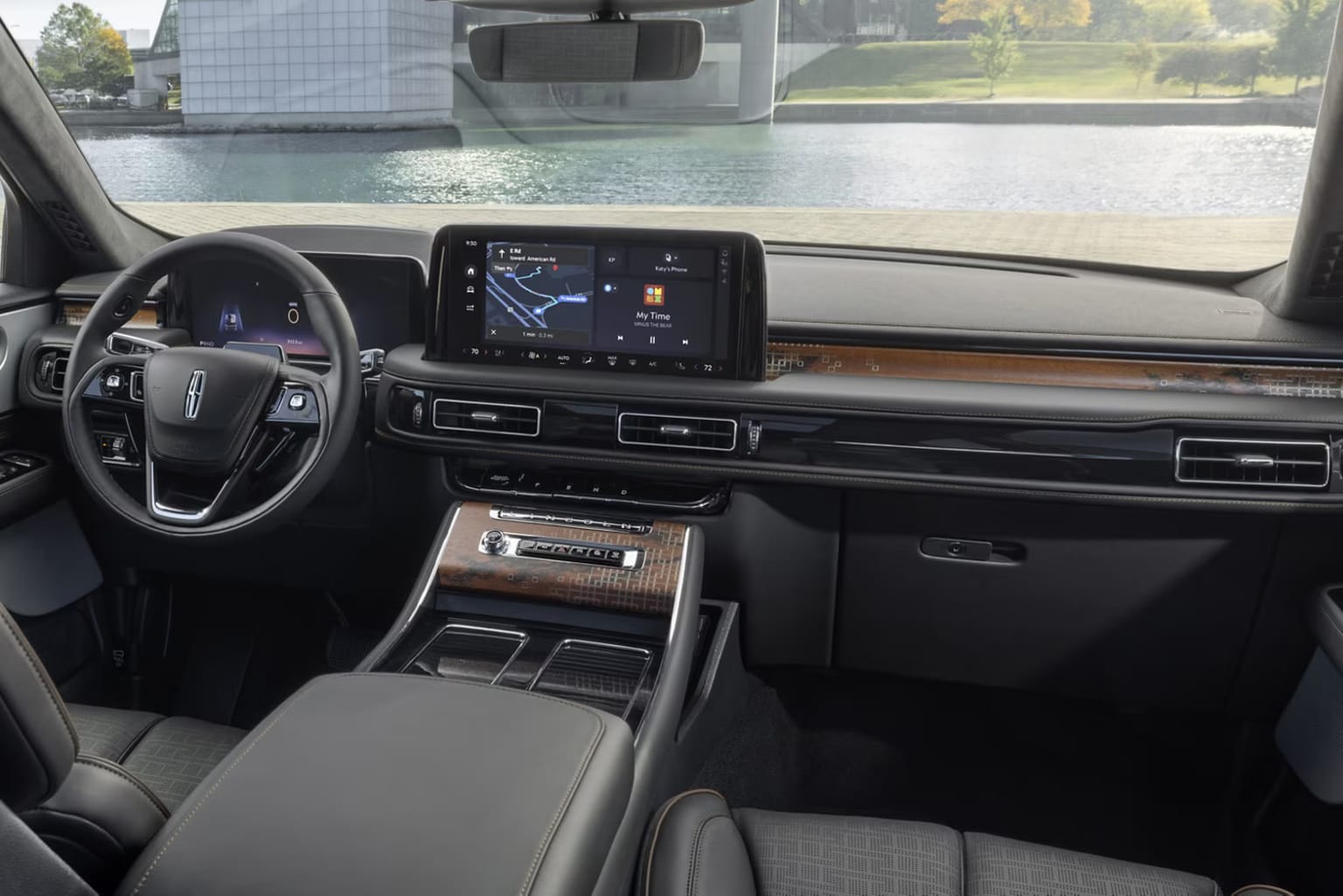 Interior of a modern vehicle with a sleek dashboard, a large touchscreen display, and a steering wheel. Background shows a body of water and a building through the windshield.