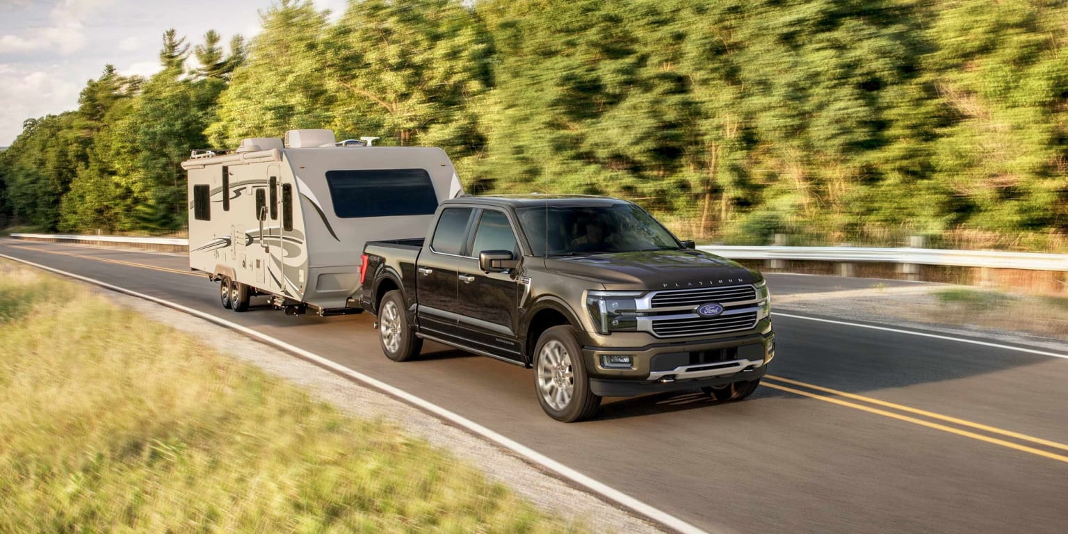 A large pickup truck towing a recreational vehicle trailer on a winding road surrounded by lush, green foliage in the background.