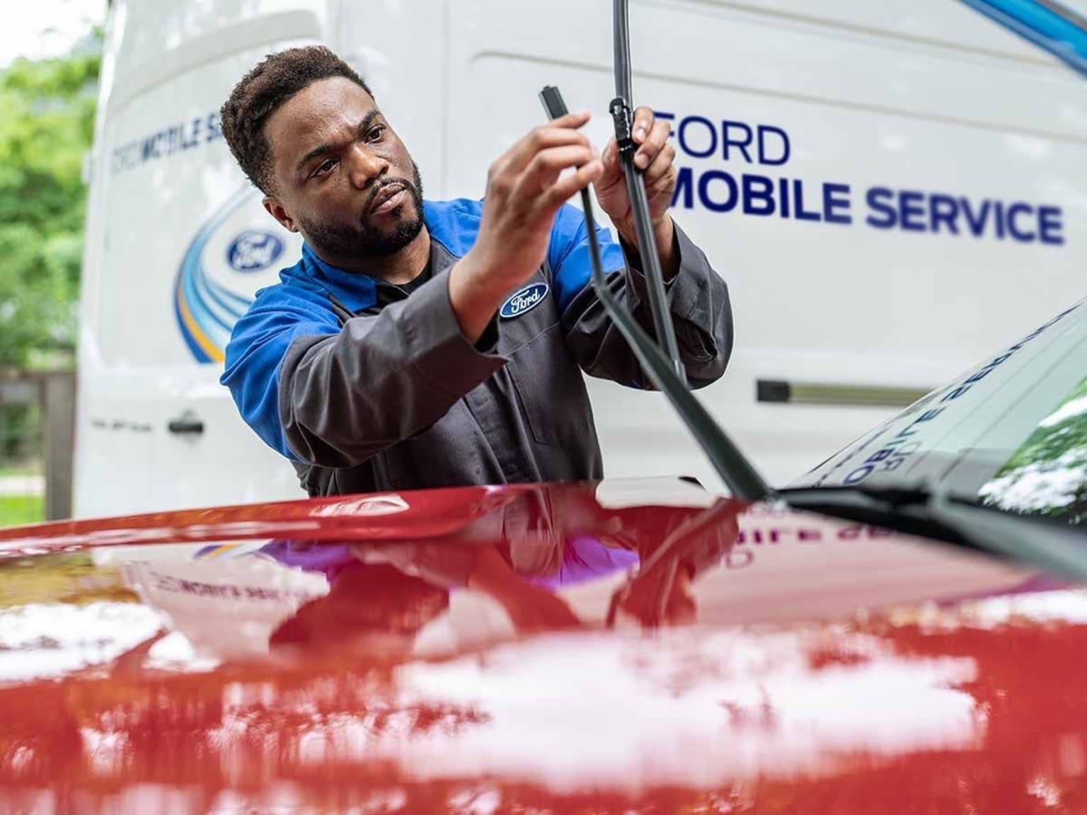 A mechanic in a blue uniform is working on the front of a service van, using tools to inspect or repair the vehicle.