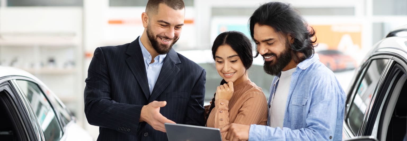 Three individuals, two men and one woman, standing together and looking at a tablet or laptop in an office setting with a glass wall or window in the background.
