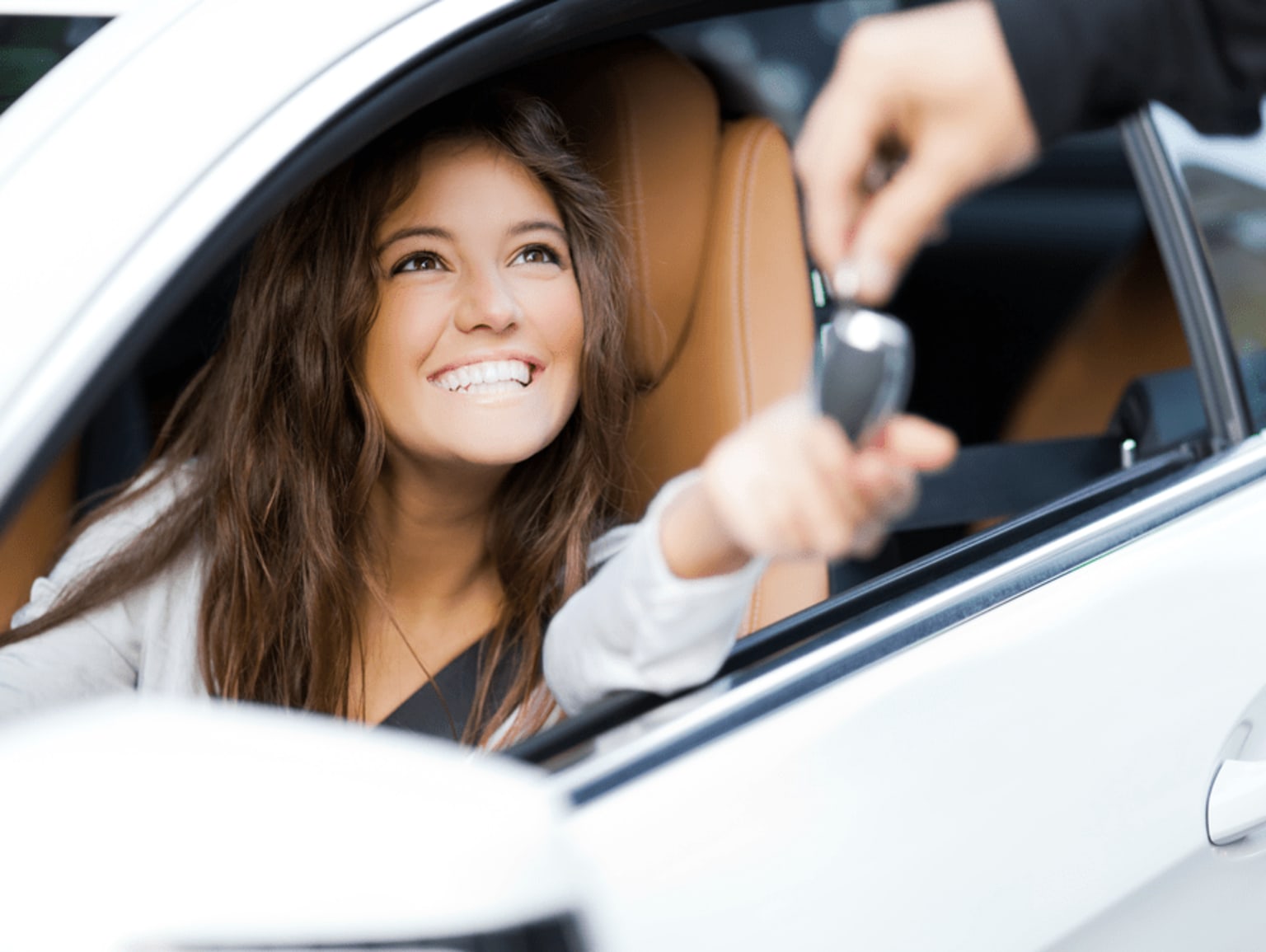 A smiling young woman with long brown hair is sitting in the driver's seat of a white car, holding the steering wheel and appearing to be driving.