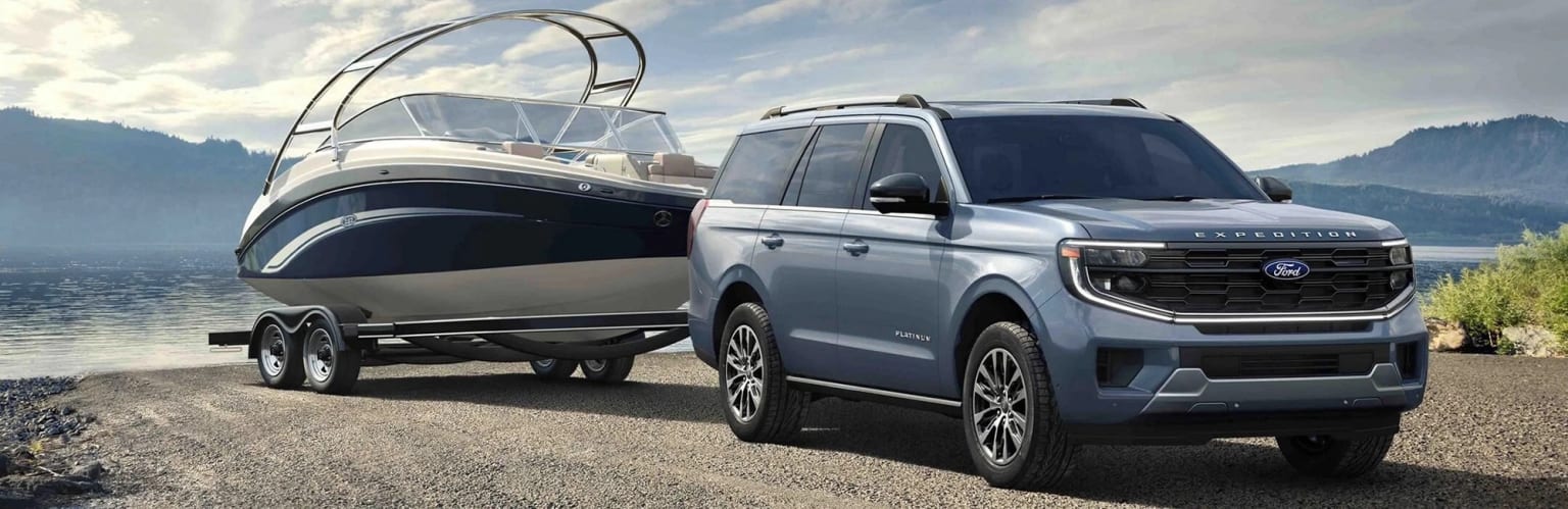 A large, silver SUV parked next to a boat trailer, under a cloudy sky and open landscape in the background.