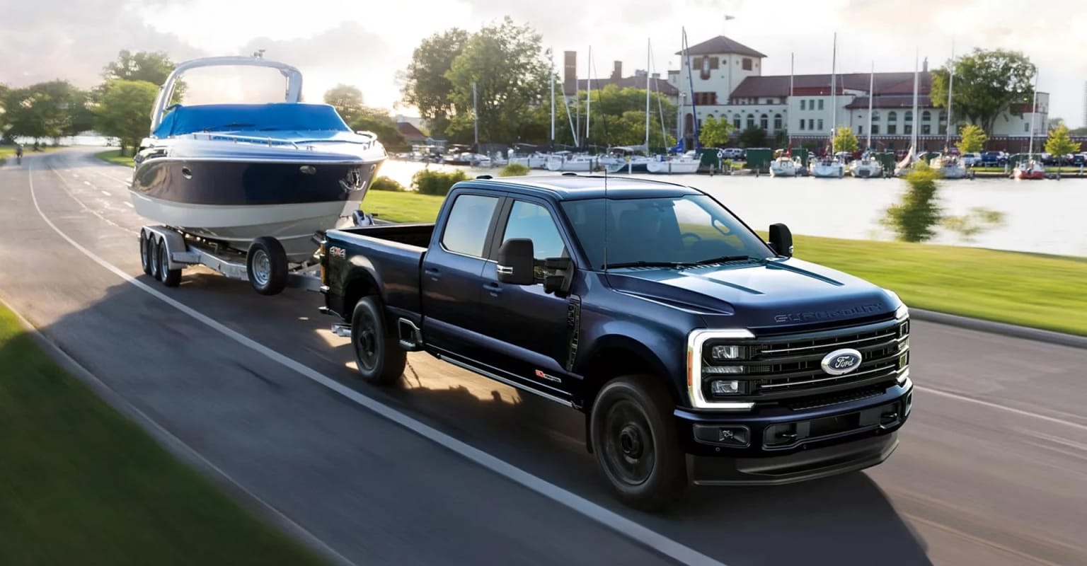 A large black pickup truck is towing a blue boat on a trailer down a road, with a town or city visible in the background.