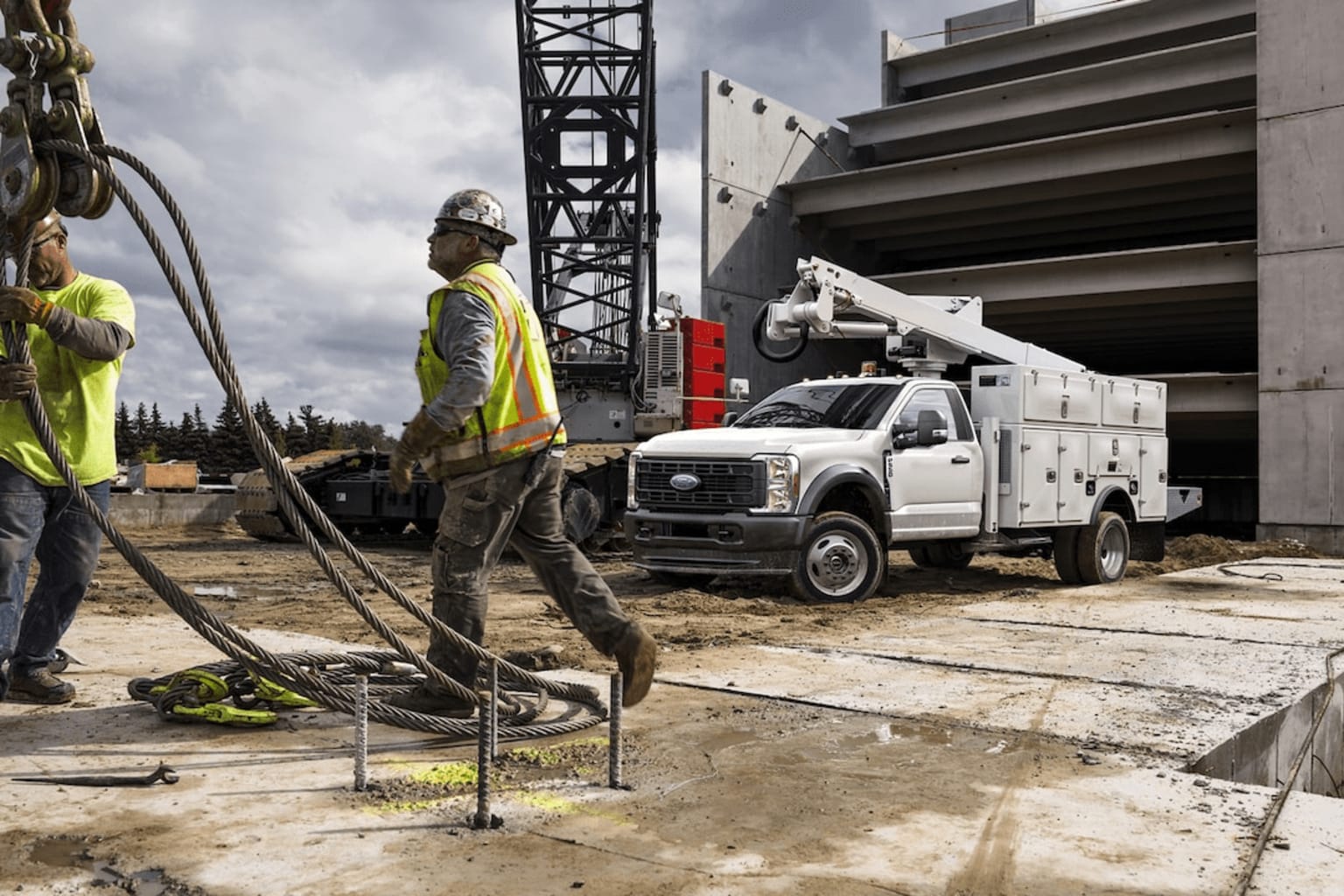 The image depicts a construction site with a worker operating heavy machinery in the foreground, and a large crane and a utility truck in the background.