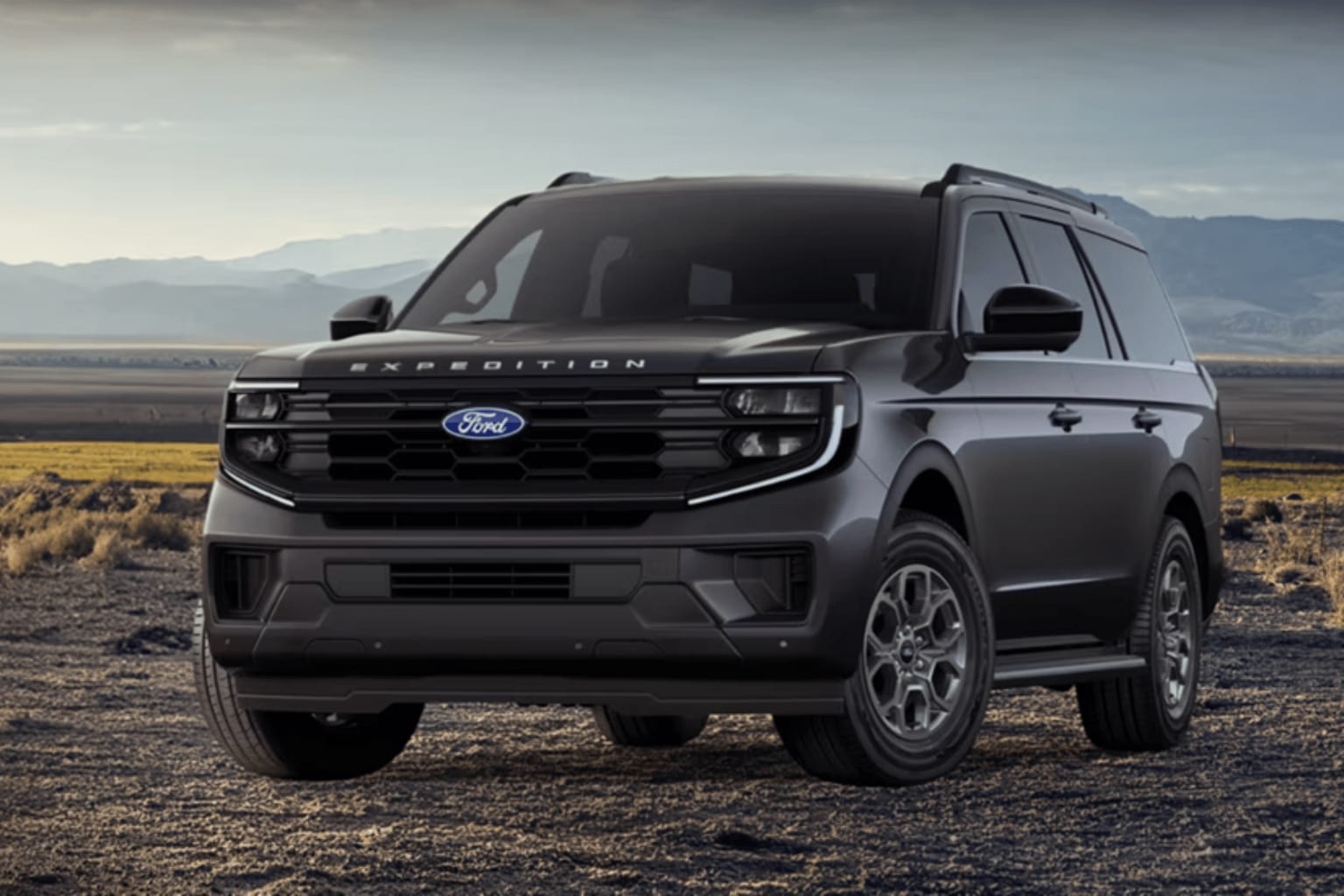 An SUV is parked on a dirt road with dry grass and distant mountains under a cloudy sky.