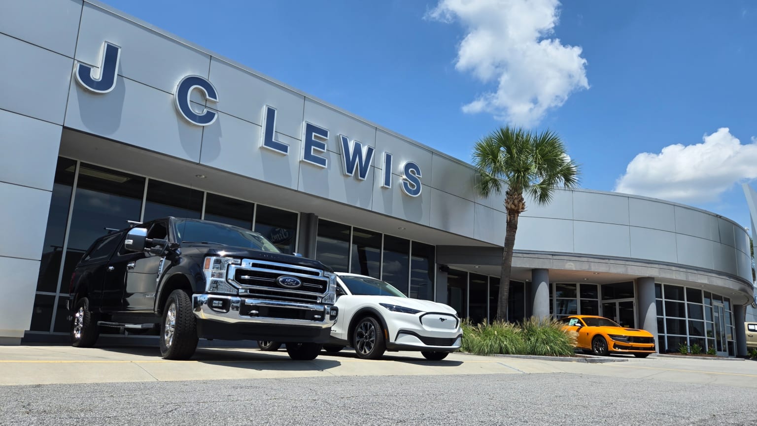 A black truck and a white electric vehicle are parked in front of a dealership with a yellow vehicle visible in the background.