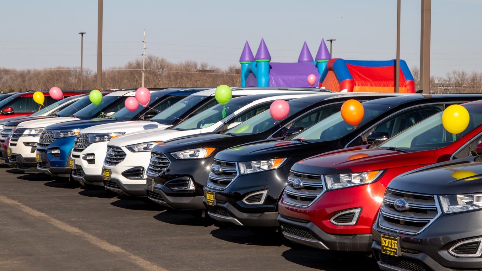 a row of cars parked in a parking lot with balloons on the top of the cars and a carnival in the background