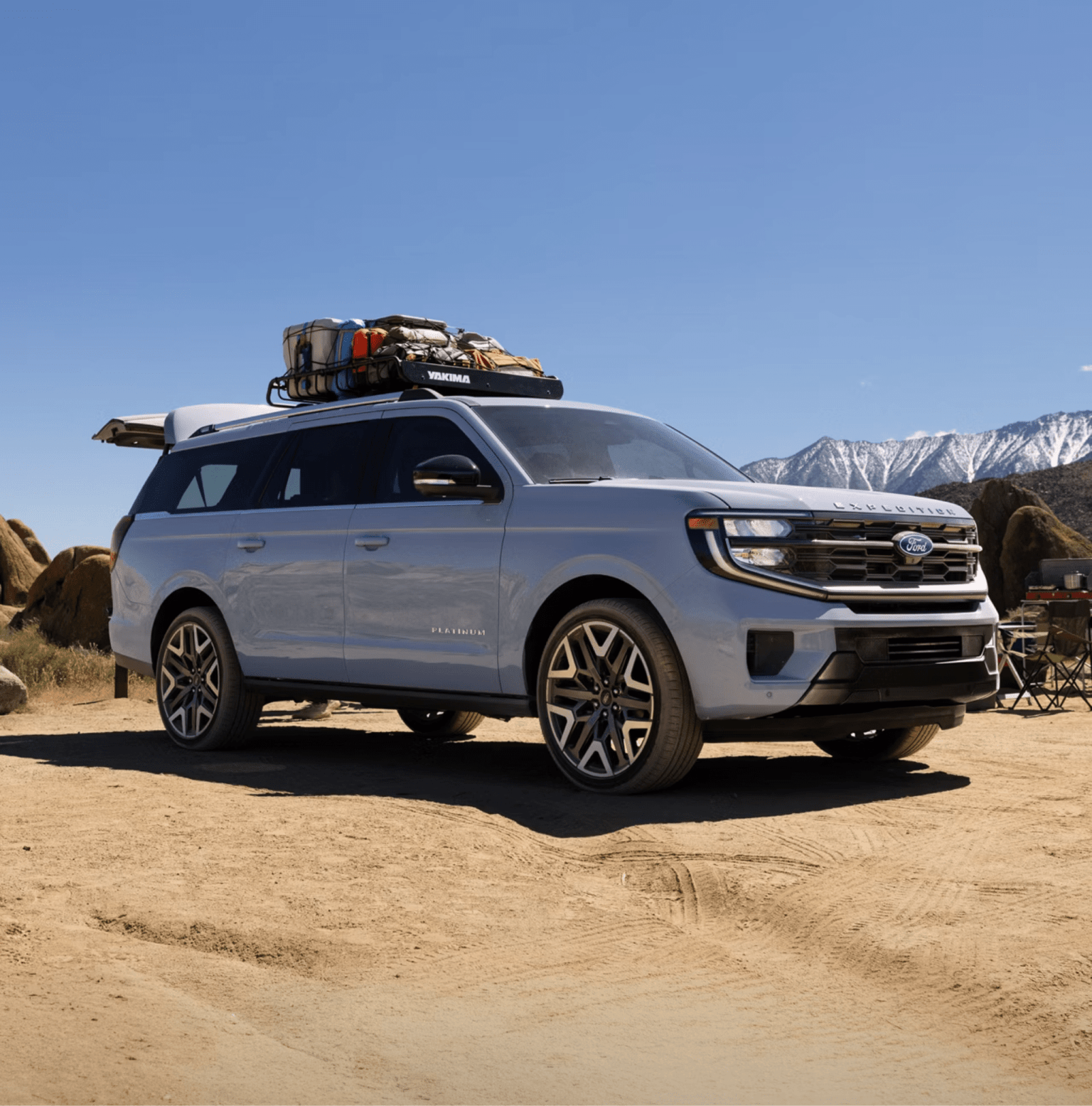 A light blue SUV with gear on its roof rack in a desert landscape with rocky hills and snow-capped mountains.