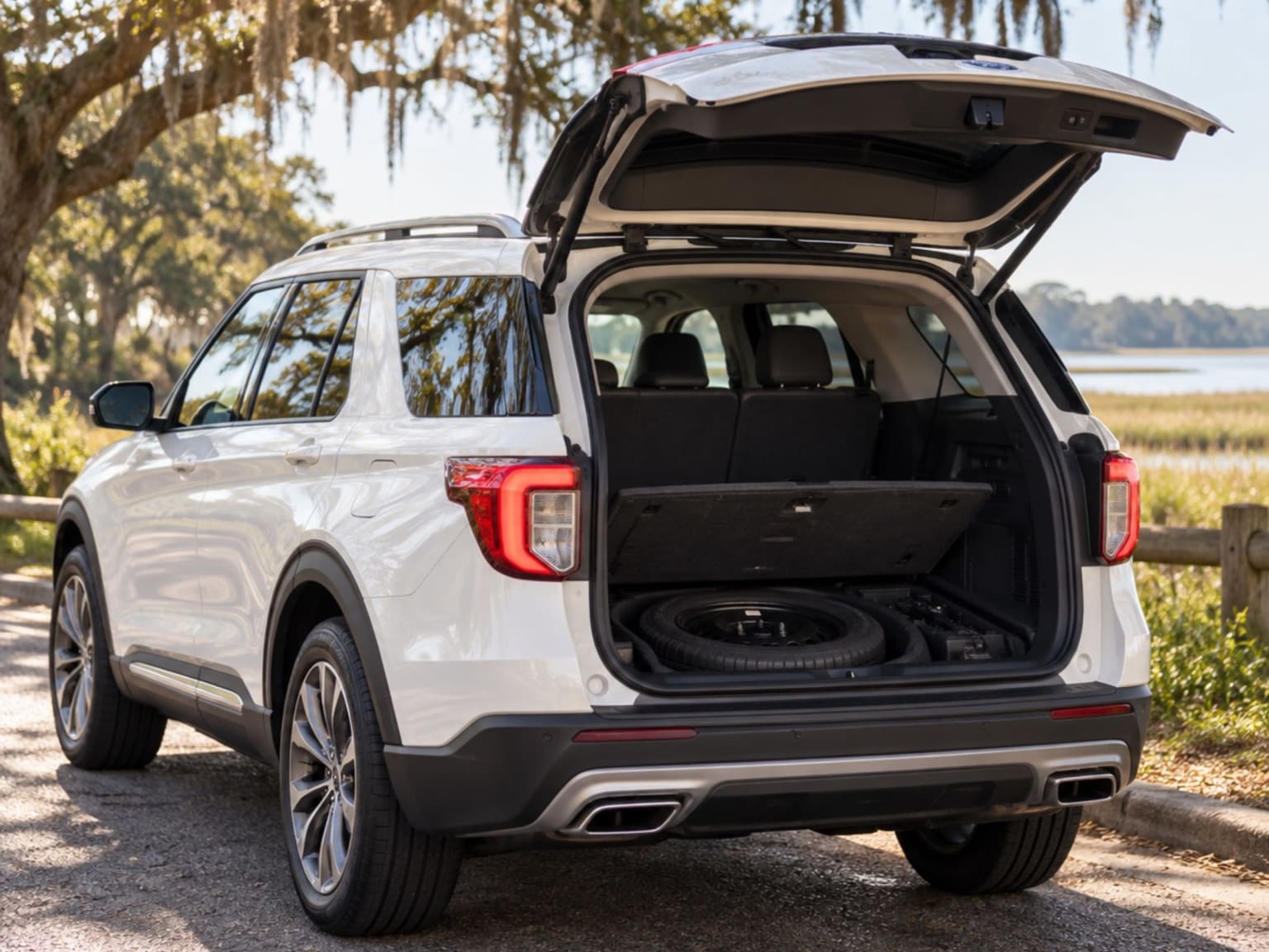 A white SUV with its trunk open on a path surrounded by trees and a body of water in the background.