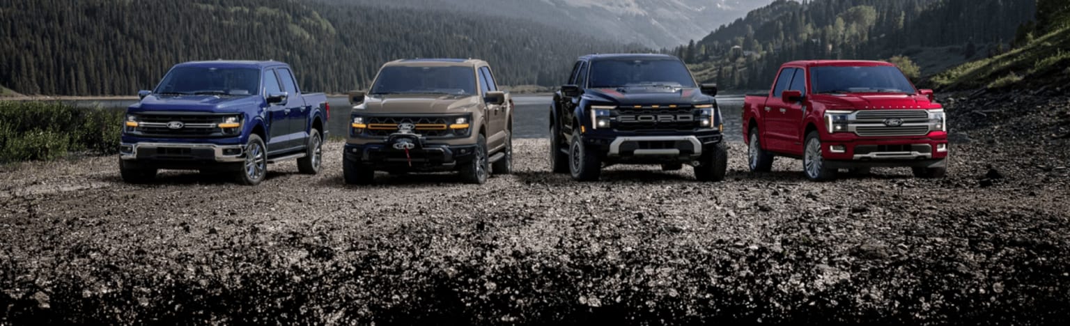 Four pickup trucks parked on a rocky, dirt road in front of a lake and a forested mountain range.