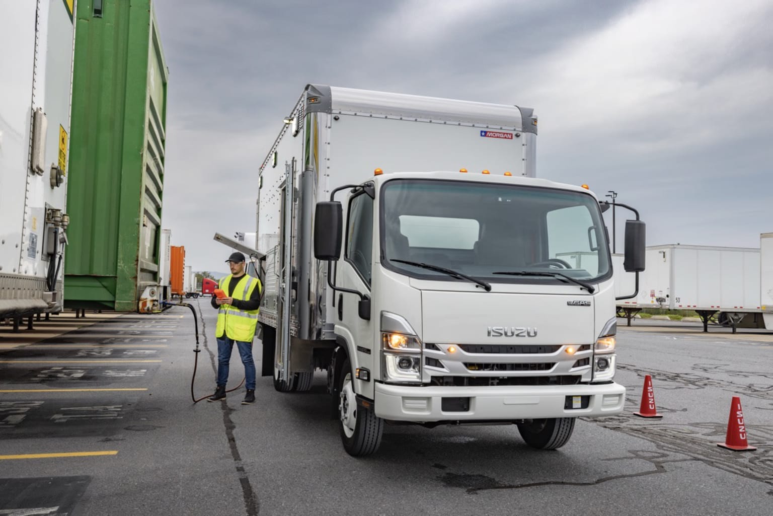 A white truck with a cargo container is parked in the foreground, while a worker in a yellow safety vest stands nearby in the background.
