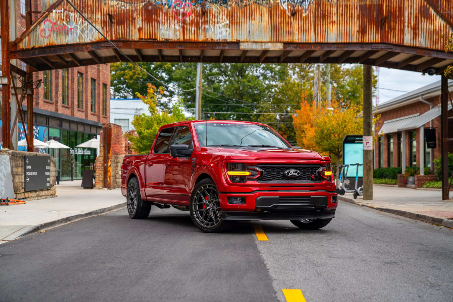 A red pickup truck parked on a street with brick buildings and trees in colorful autumn foliage.