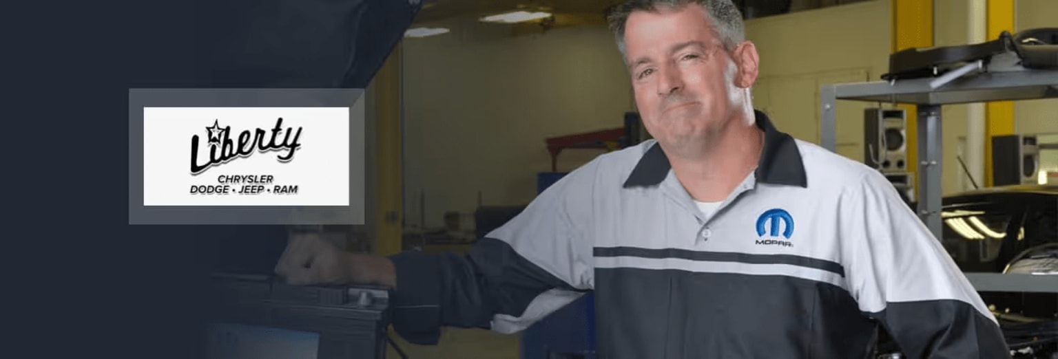The image shows a middle-aged man wearing a white and gray polo shirt, standing in front of a dealership sign.