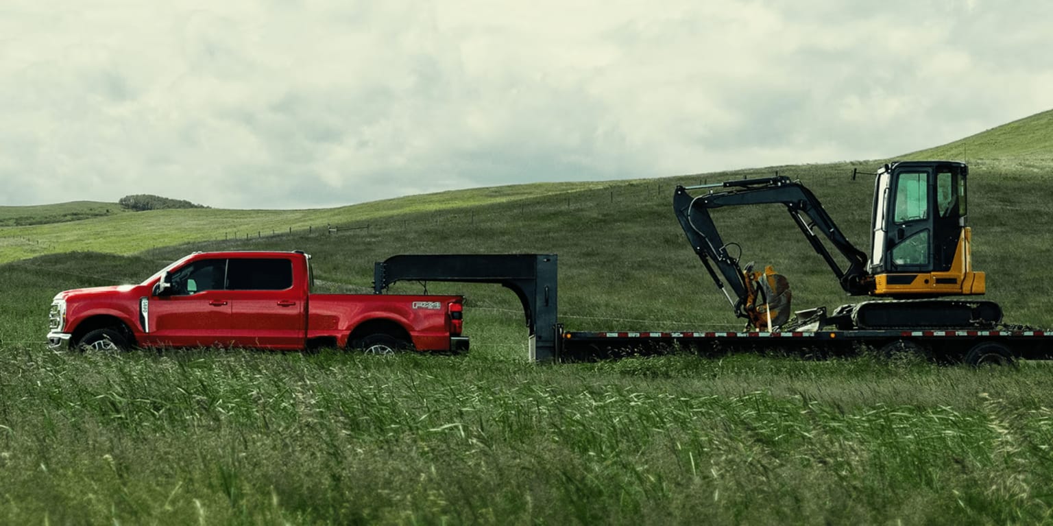 A red pickup truck towing a yellow excavator on a flatbed trailer in a grassy field with green hills and cloudy sky behind.