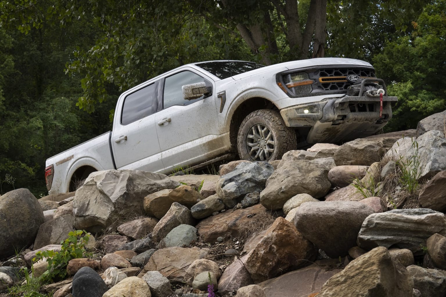 A white pickup truck off-roading on rocky terrain with lush greenery and trees in the background.
