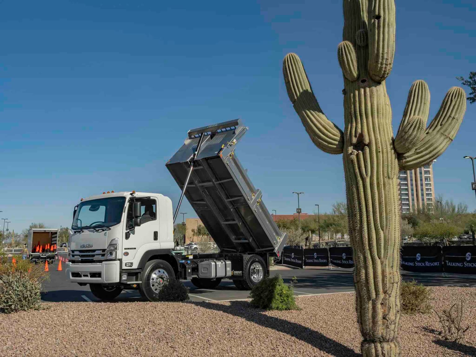 A white dump truck with its bed raised next to a large saguaro cactus, with a modern building and clear blue sky in the background.
