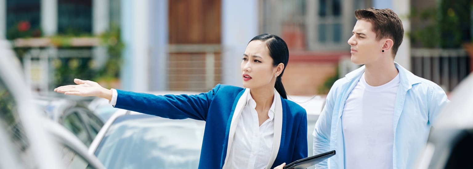 A woman in a blue blazer gestures towards a car while a man in a light blue shirt looks on, with buildings in the background.
