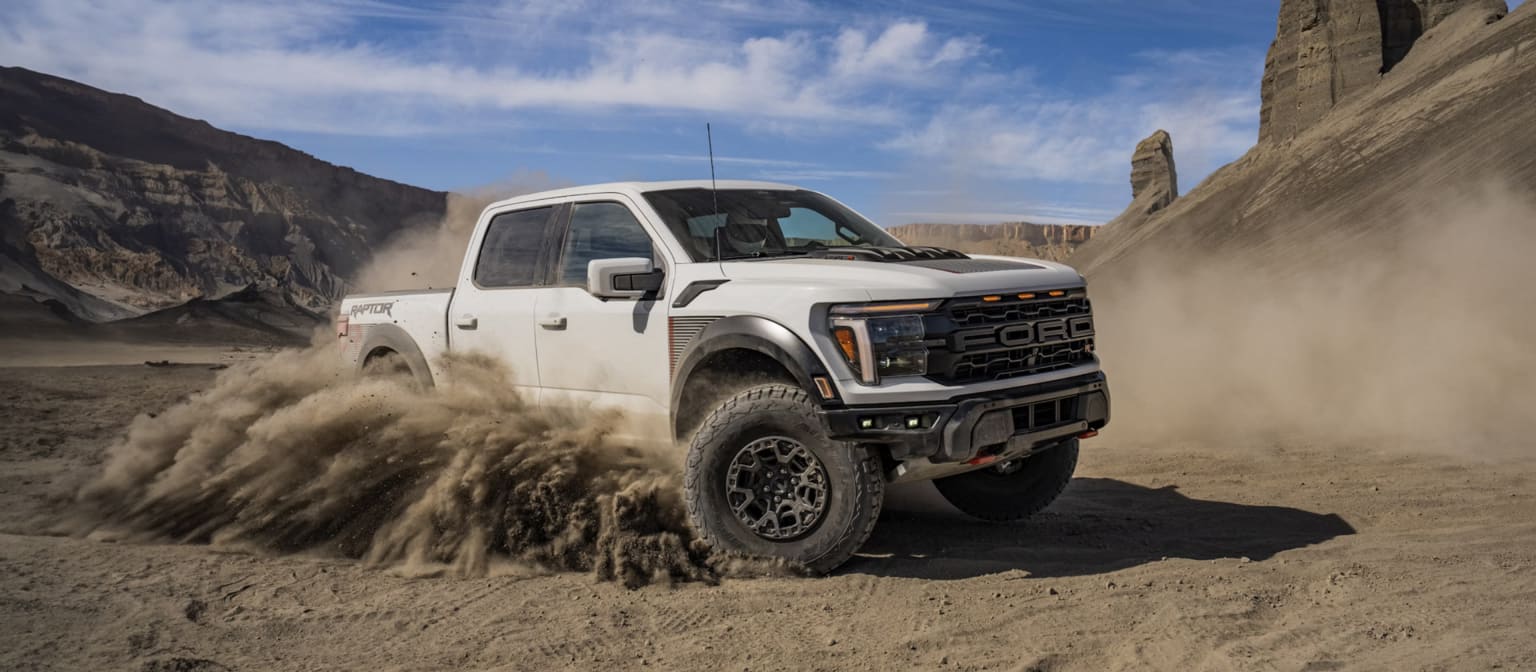 A rugged, off-road pickup truck kicking up a cloud of dust as it navigates through a desert landscape with towering cliffs in the background.