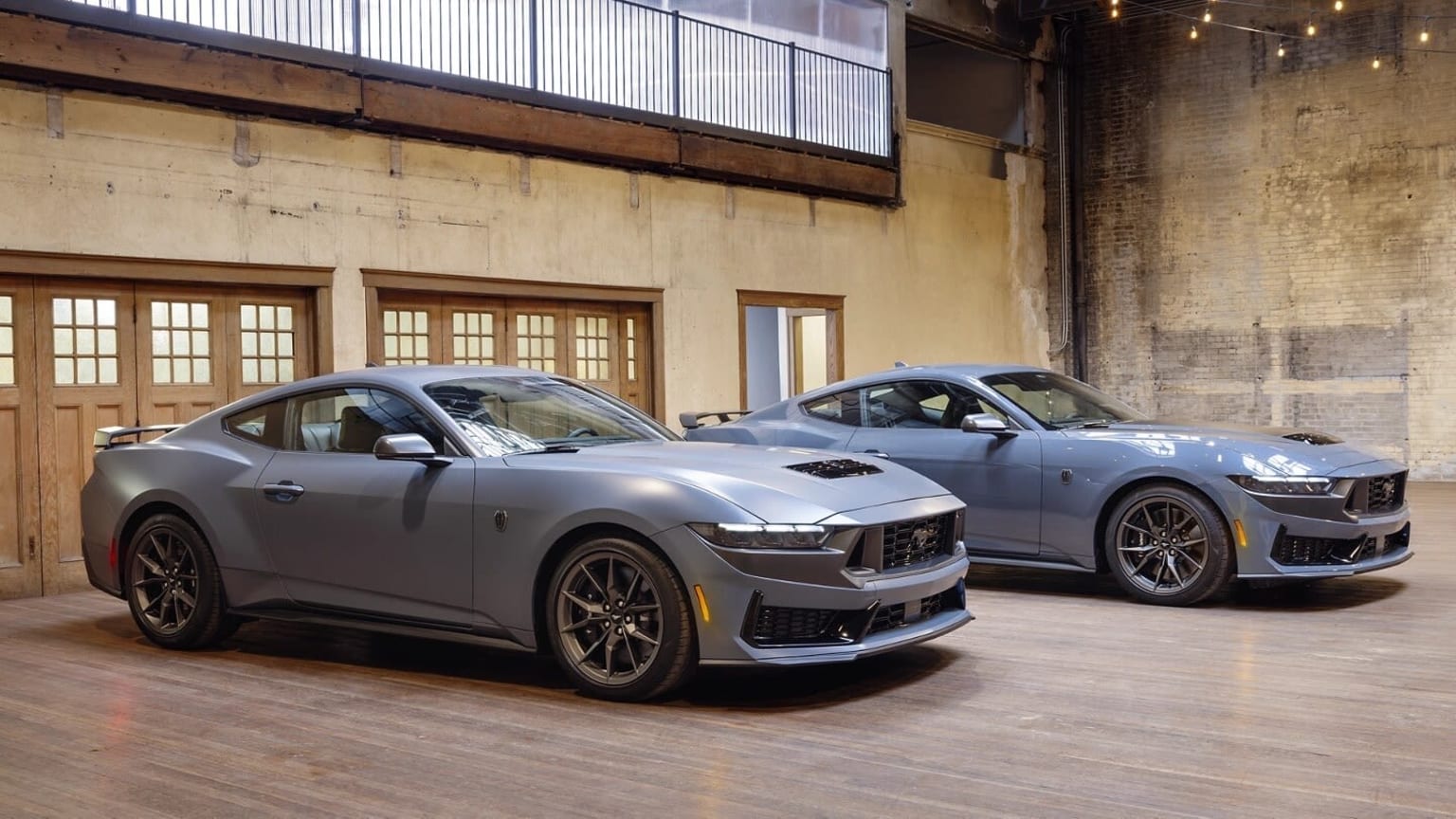 Two high-performance sports cars, one silver and one gray, are parked in a dimly lit industrial-style garage with brick walls and large windows.