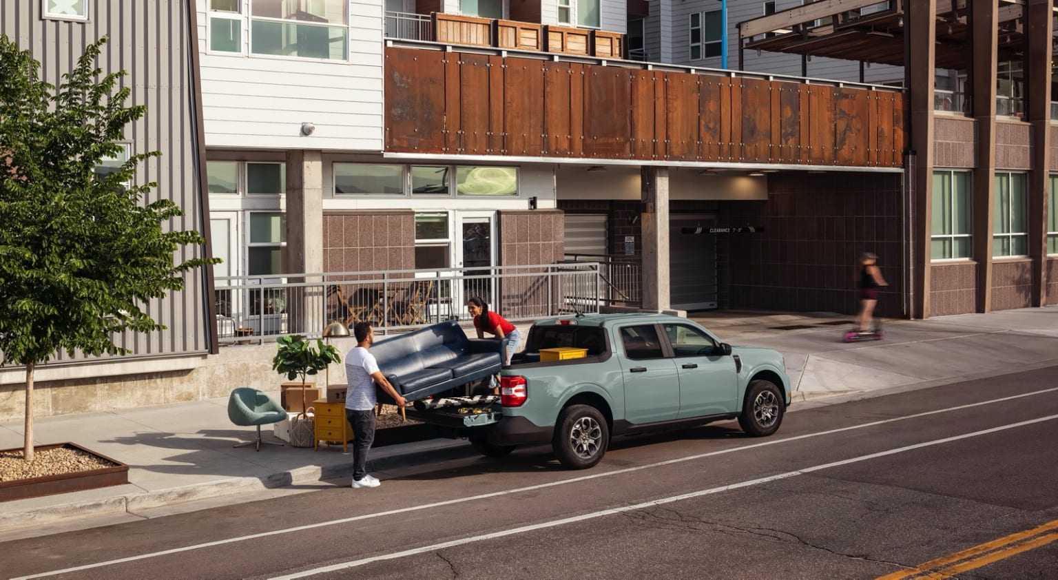A pickup truck parked on the street in front of a multi-story building with wooden balconies, and a person walking in the background.