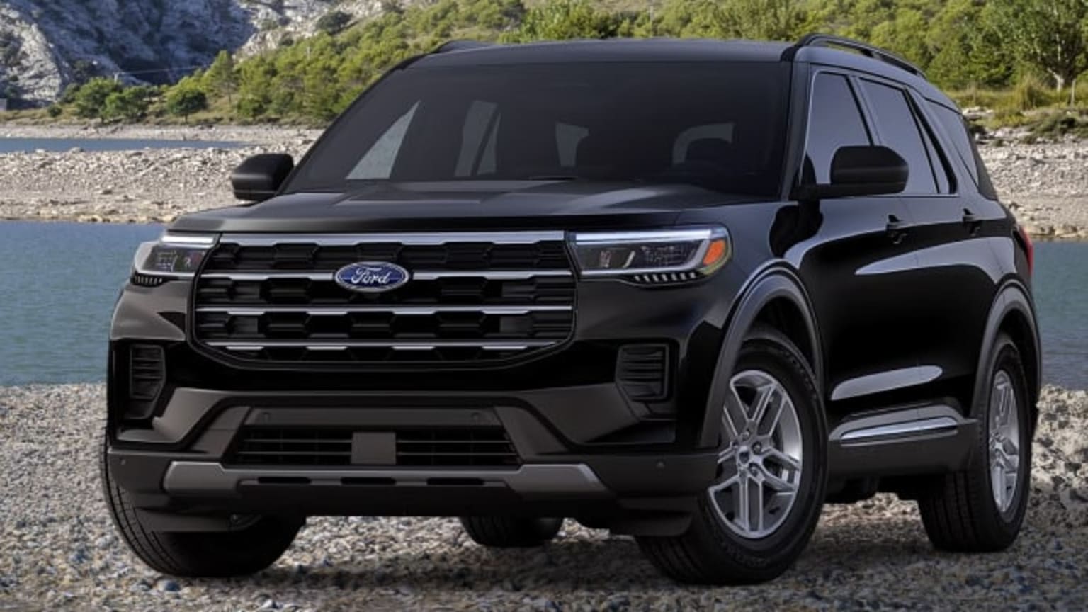 A black SUV is parked on a rocky shore with a body of water and a wooded hillside in the background.