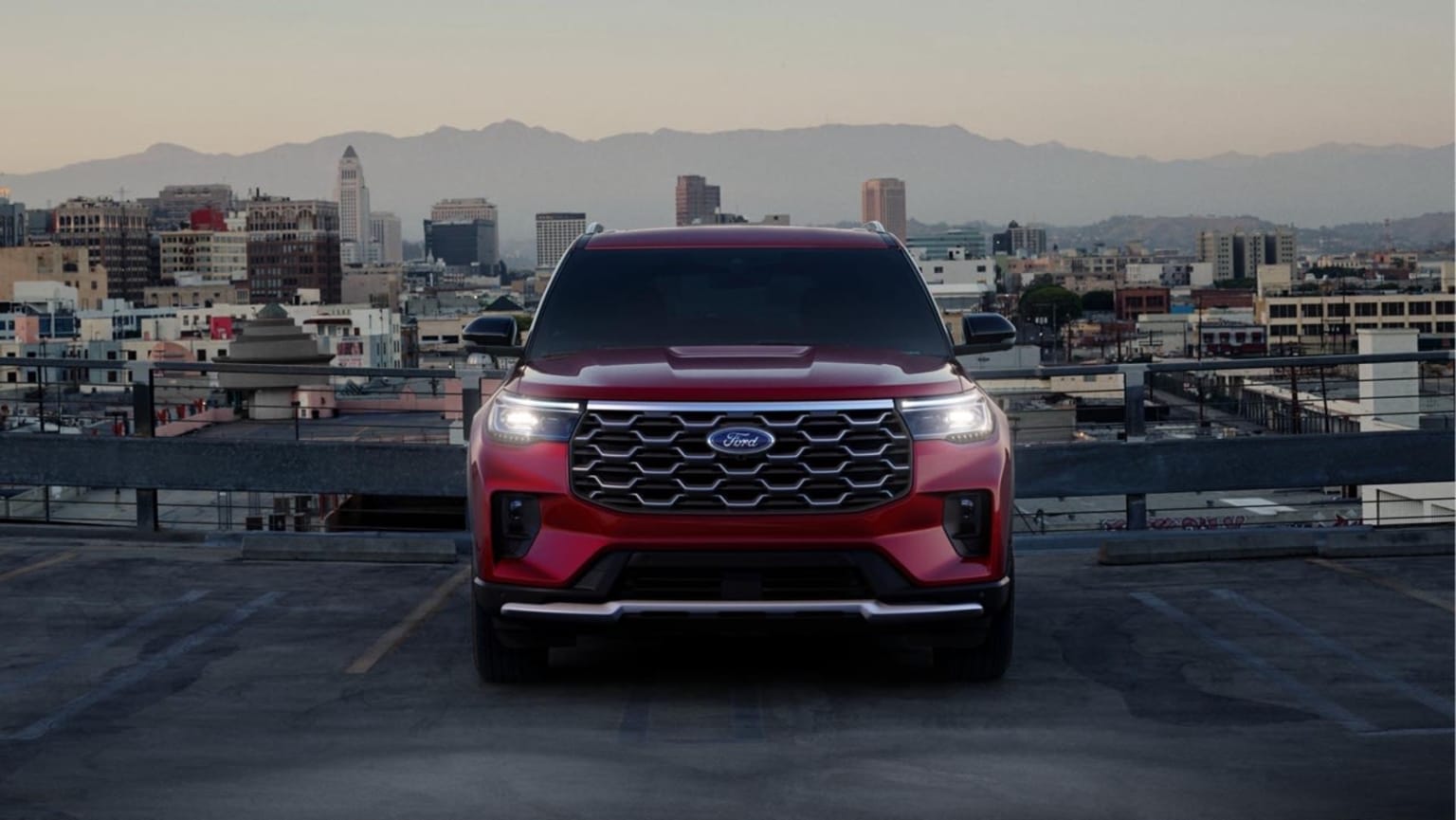 A red SUV with a distinctive grille design in the foreground, cityscape with skyscrapers and mountains in the background.
