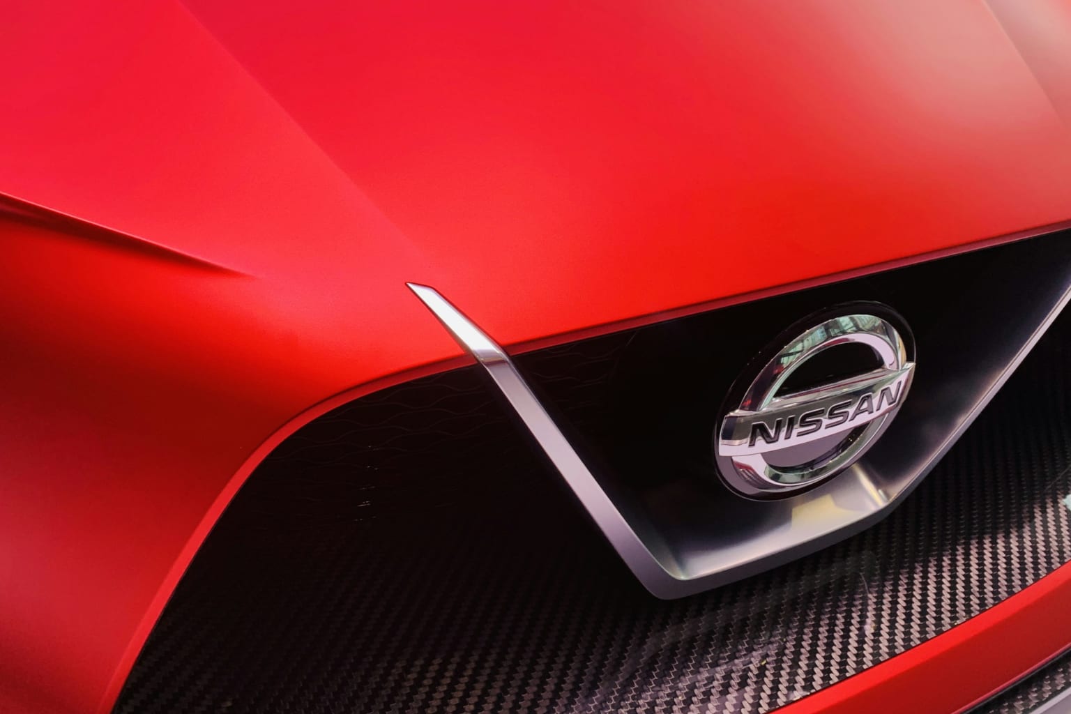 The image shows the front end of a red sports car, with the logo prominently displayed on the grille against a dark, carbon fiber-like background.