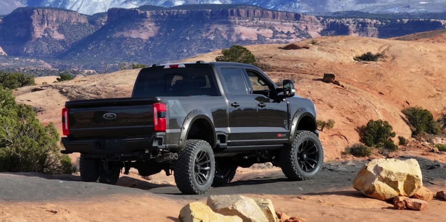 Off-road vehicle navigating rocky, mountainous terrain with towering cliffs and vast, arid landscape in the background.