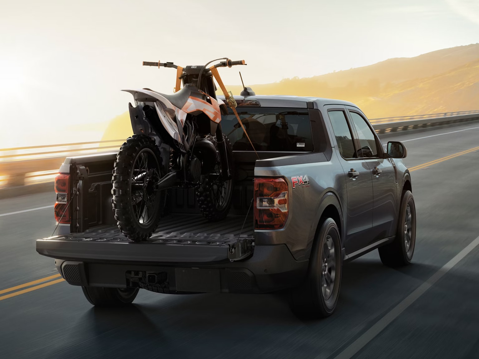 A dirt bike being loaded into the bed of a pickup truck on a highway with a sunset in the background.