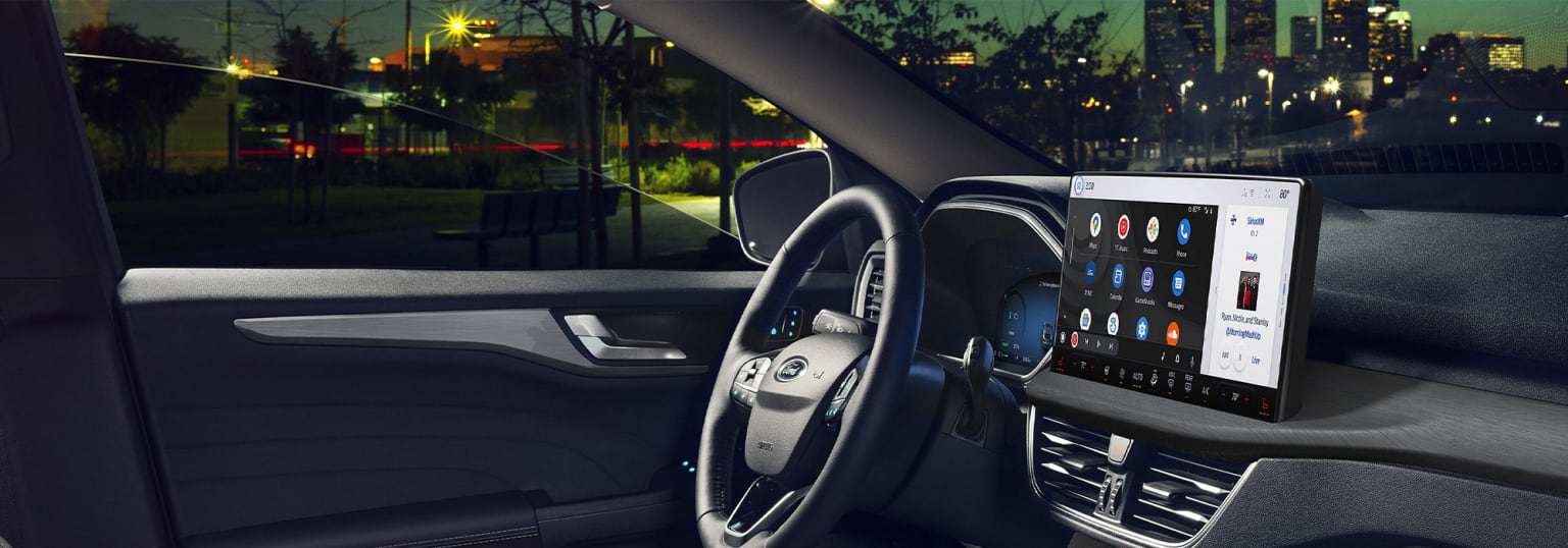 The image shows the interior of a modern car at night, with the steering wheel, dashboard, and a large touchscreen display visible in the foreground, while the background depicts a dimly lit urban landscape outside the vehicle.
