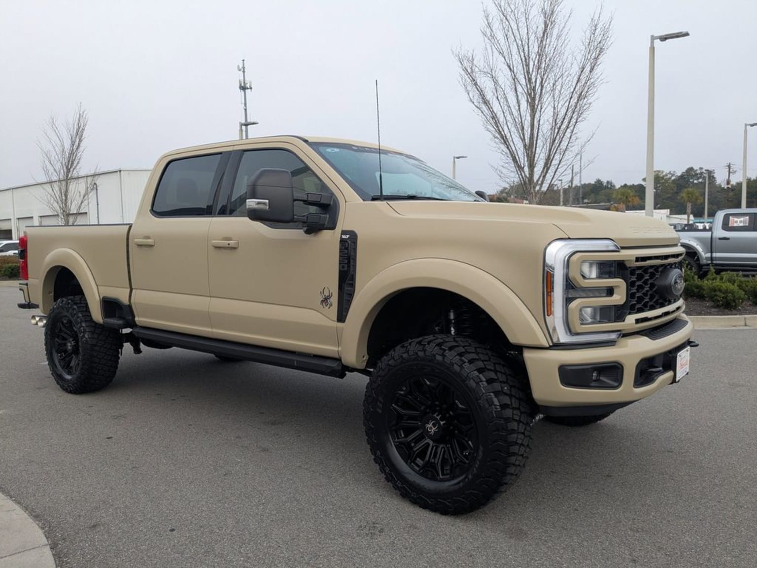 A lifted tan pickup truck with large black wheels in a parking lot with a building and trees in the background.