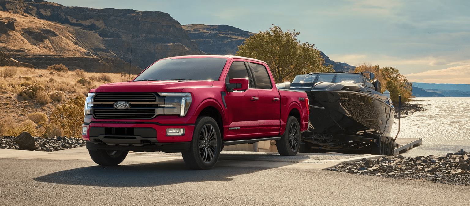 A red truck towing a boat on a trailer near a rocky shore and a calm body of water, with arid hills in the background.