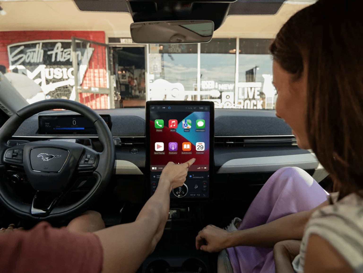 Interior of a vehicle with the driver's hands on the steering wheel and a smartphone displaying various app icons on the dashboard.