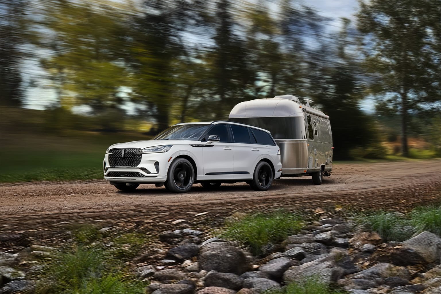 An image of a large white SUV towing a silver Airstream trailer on a dirt road surrounded by trees and foliage.