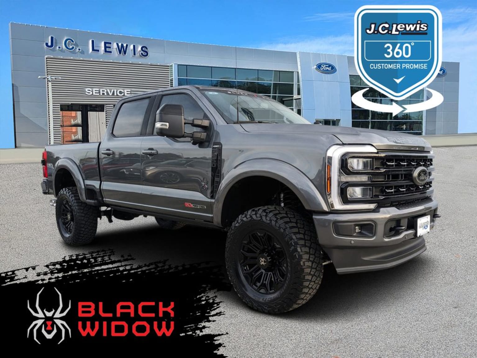 A large, lifted pickup truck is prominently displayed in the foreground, with an automotive dealership building visible in the background against a clear blue sky.