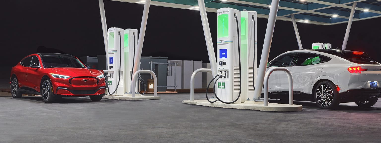 two electric cars at a charging station with a red car in the foreground and a white car in the background