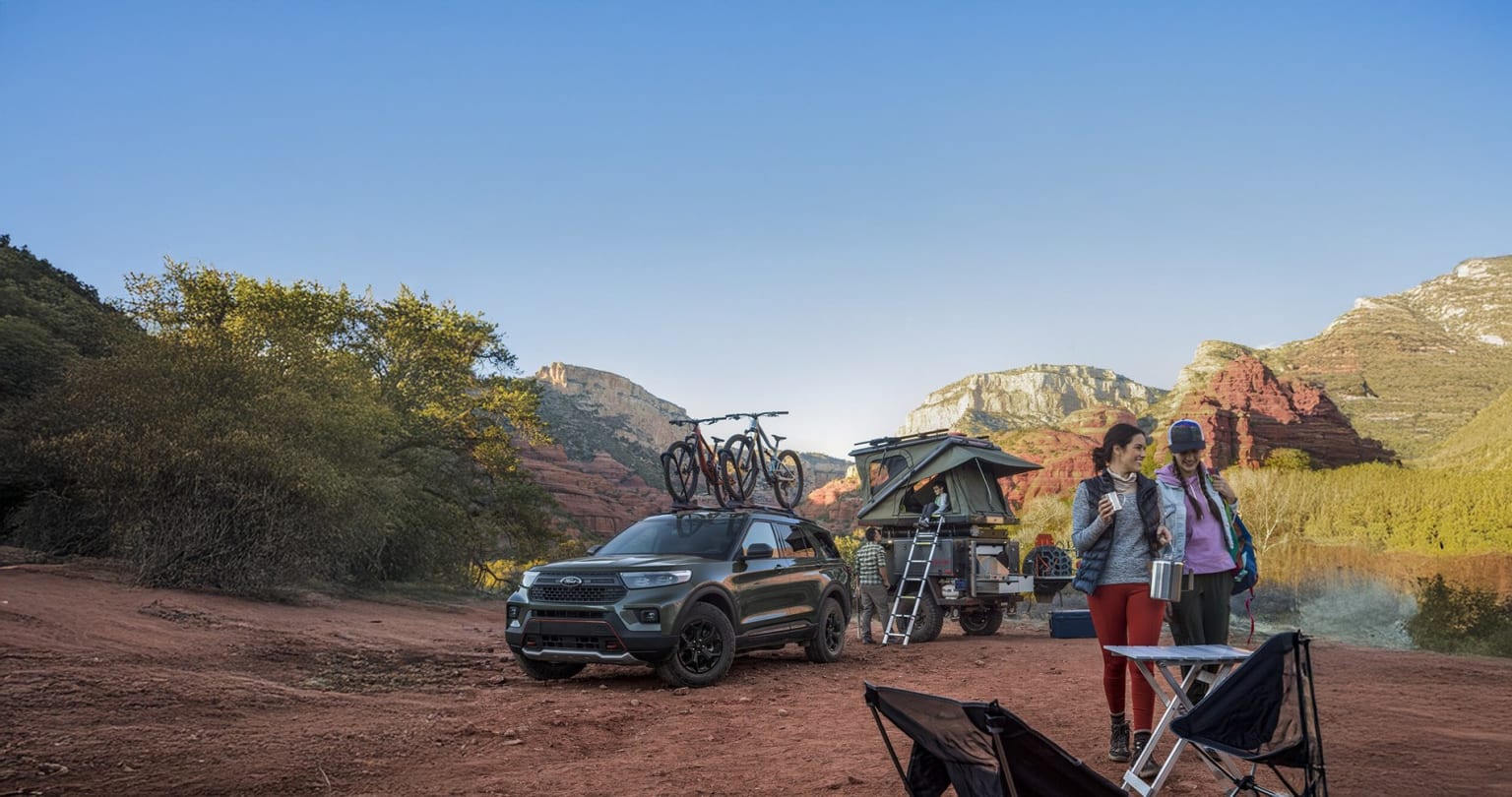 Off-road vehicle with rooftop tent and bicycle rack parked on dirt road surrounded by red rock formations and green vegetation in desert landscape.