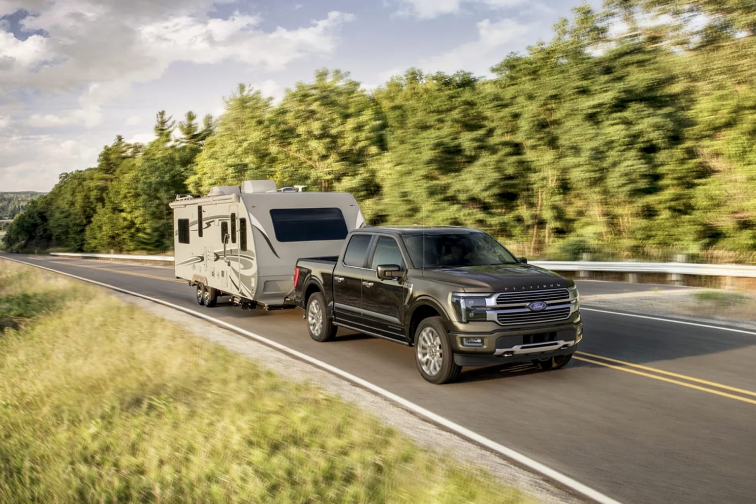 A dark pickup truck towing a travel trailer on a paved road with a blurred background of green trees and a cloudy sky.
