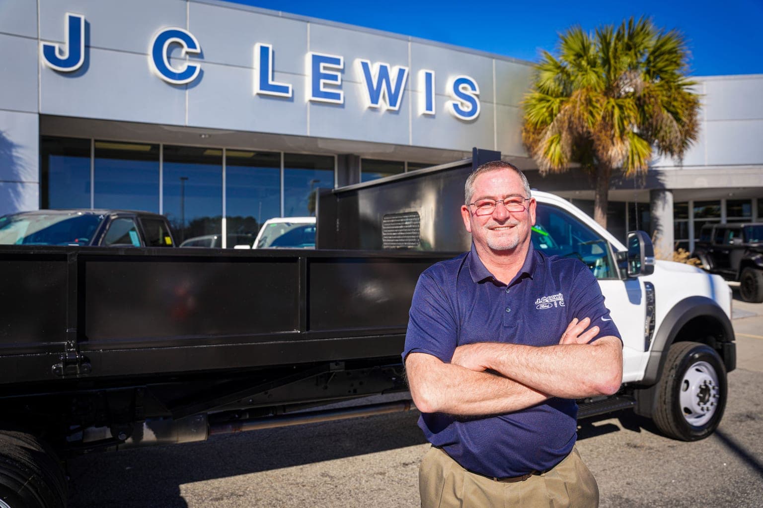 The image shows a man in a blue uniform standing in front of a large truck with a dealership logo visible on the building behind him.