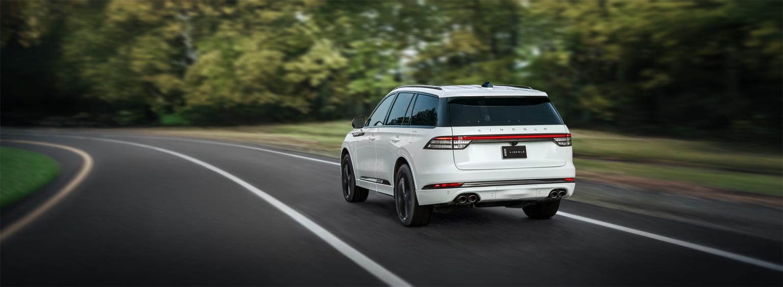 A white SUV driving on a road with lush green foliage in the background.