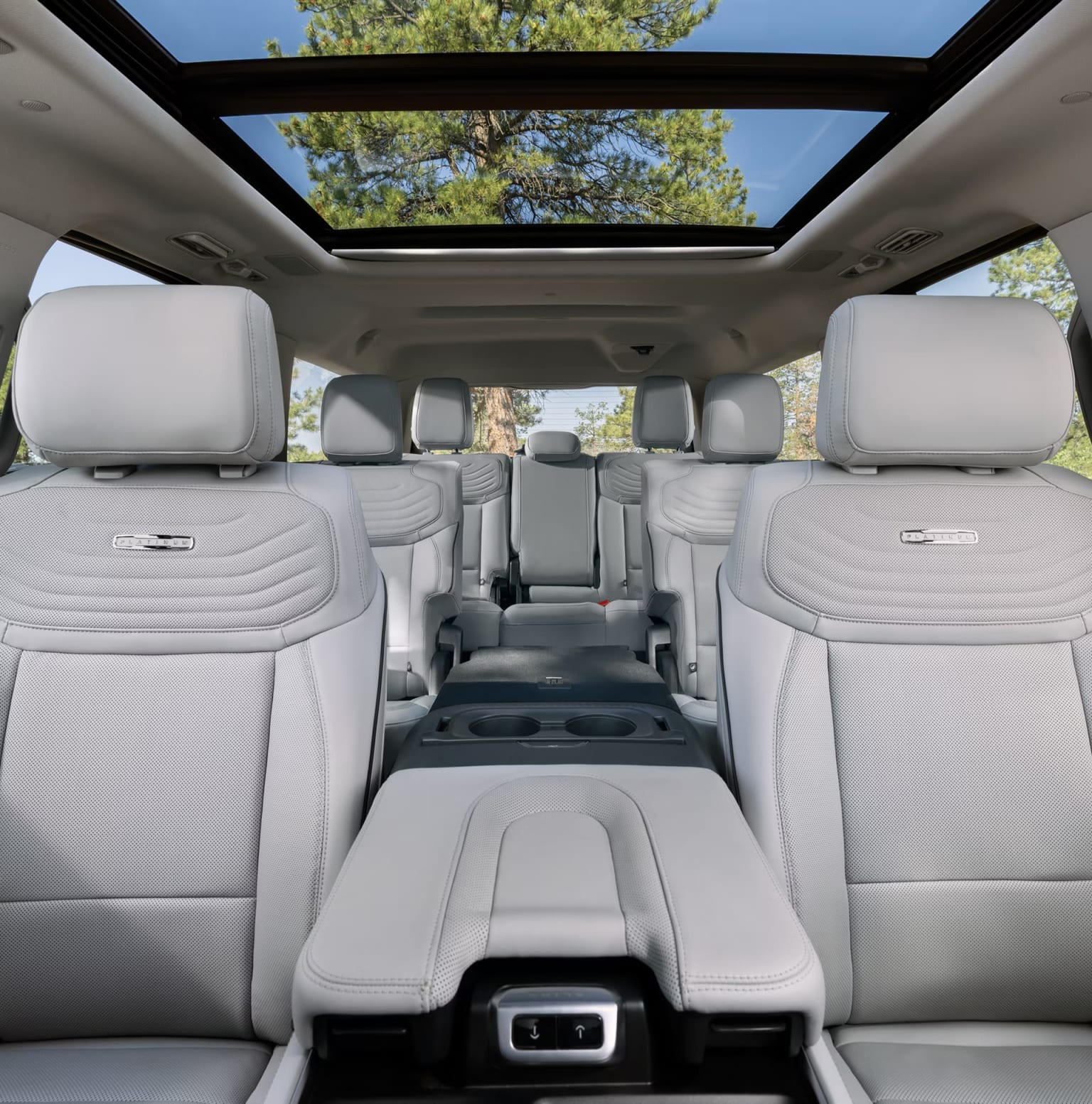 Interior of a spacious vehicle with rows of comfortable white seats and a panoramic sunroof overlooking trees and sky.
