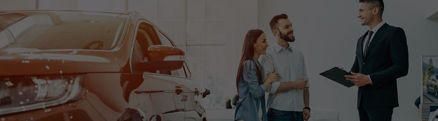 A couple standing in front of a vintage-style car, the man with a beard and the woman with long hair, both dressed casually and appearing to be in a relaxed, affectionate moment.