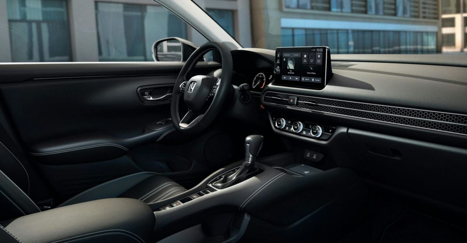 A photo of a modern car's interior, showcasing a sleek dashboard with a touchscreen and gear shifter in the foreground, set against a blurred urban backdrop.