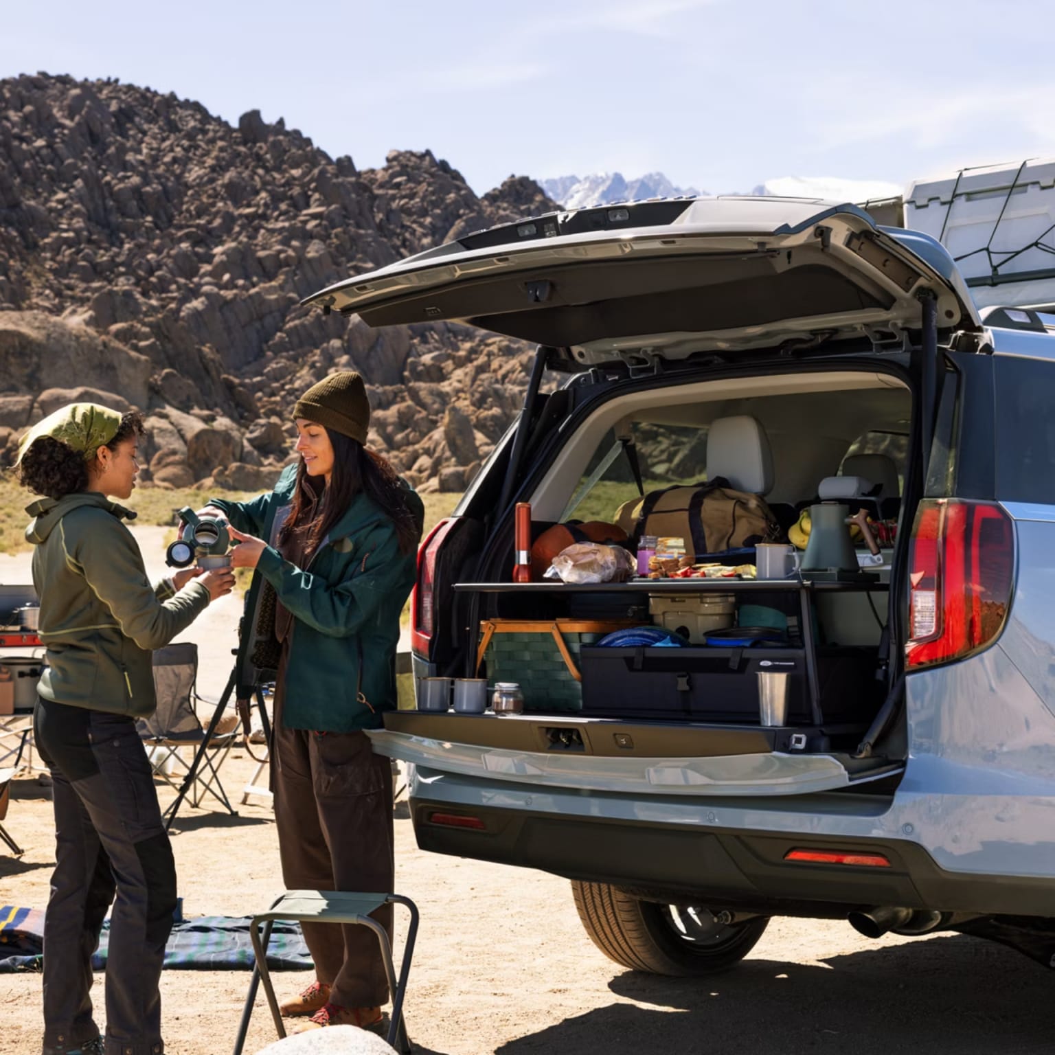 Two women are packing camping gear from the open trunk of a SUV, with a rocky, mountainous landscape in the background.