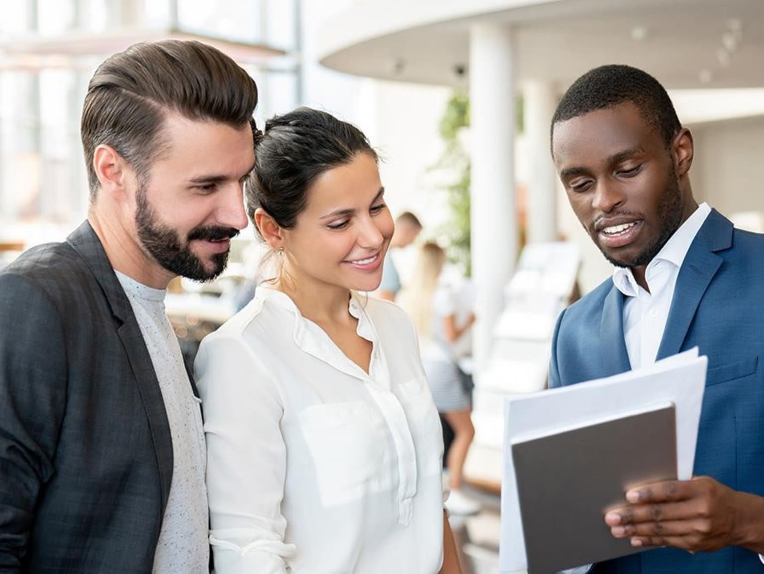 Three individuals, two men and one woman, are engaged in a conversation while holding documents in a professional setting with a bright, open background.