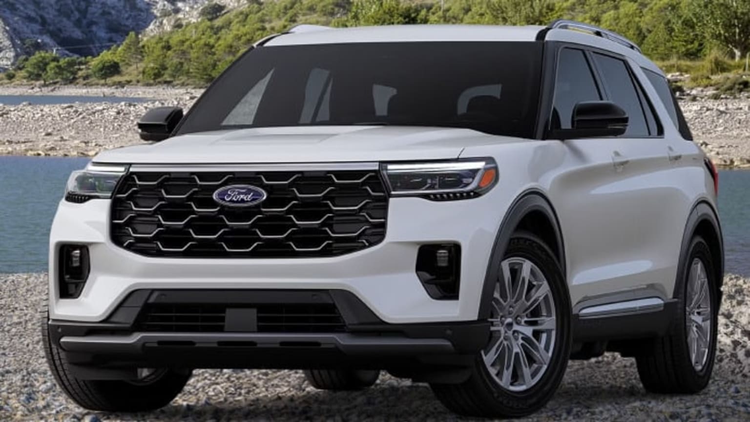 An SUV is parked on a rocky shore with a body of water and a wooded hillside in the background.