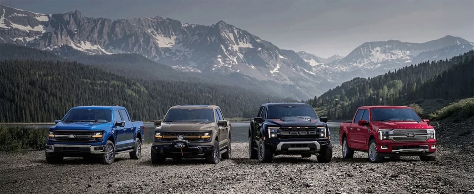 Four pickup trucks in different colors parked in a remote mountainous landscape with snow-capped peaks in the background.