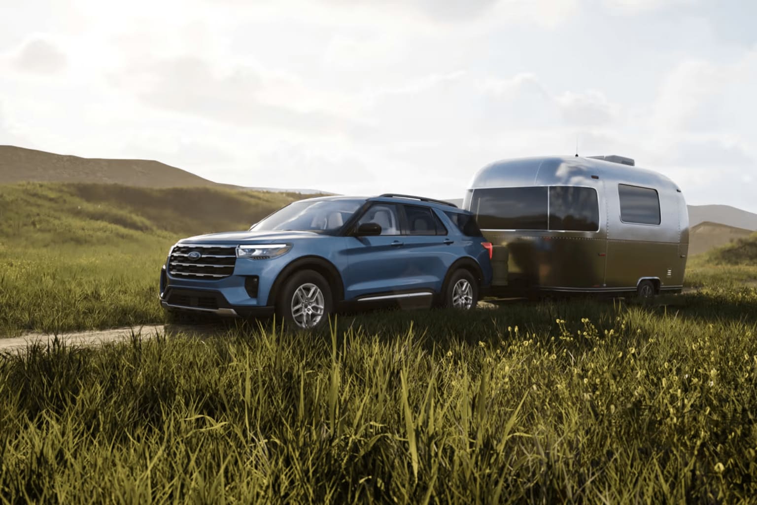 Blue SUV towing a silver Airstream trailer through a grassy field with rolling hills in the background.